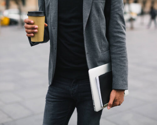 man walking in urban area with laptop, spiral notebook and coffee